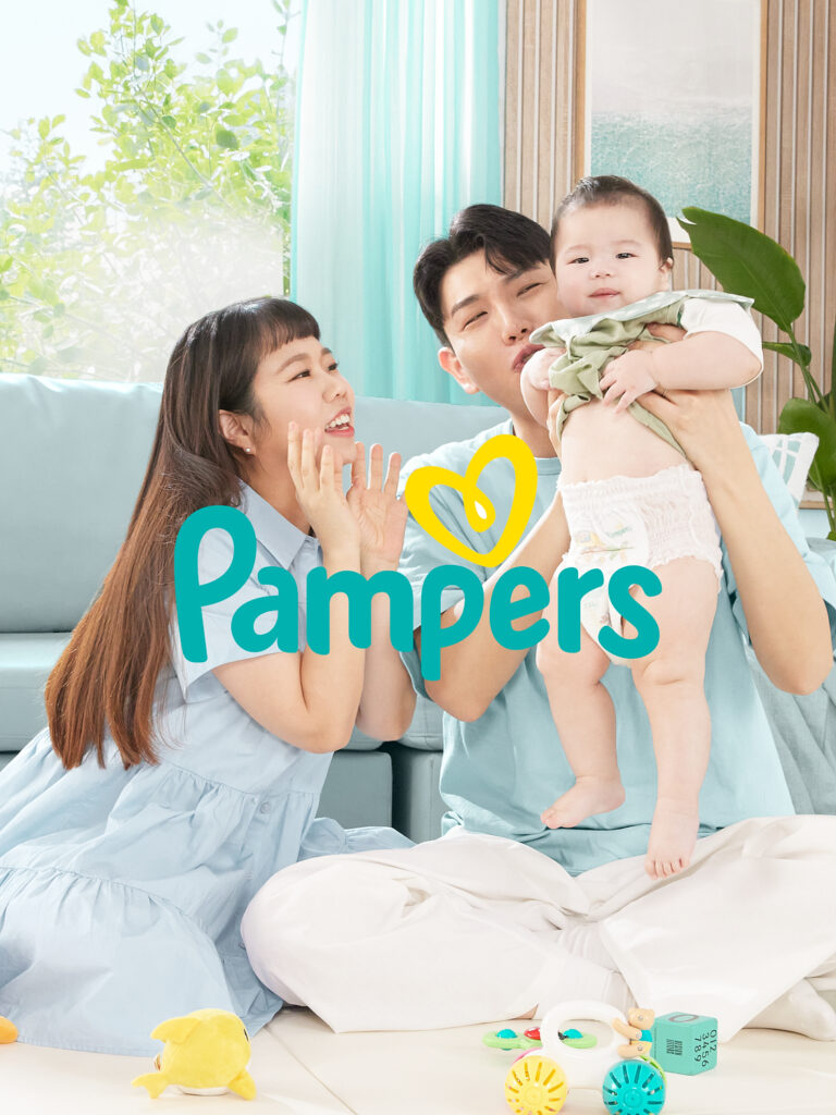 팸퍼스 브랜드 캠페인 광고 촬영. 민트 톤 거실에서 아빠가 아기를 들어 뽀뽀하고 엄마가 웃으며 바라보는 가족 씬 세로 메인 컷.